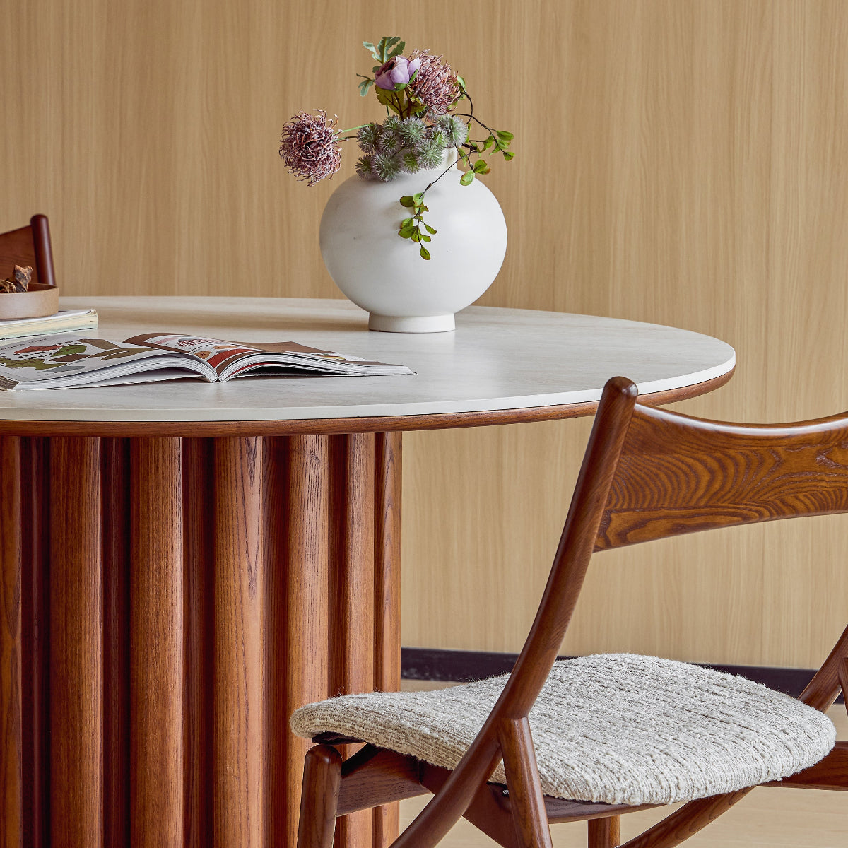 Round table with a vase of flowers and a book, wooden chair with a cushioned seat, against a wooden wall.