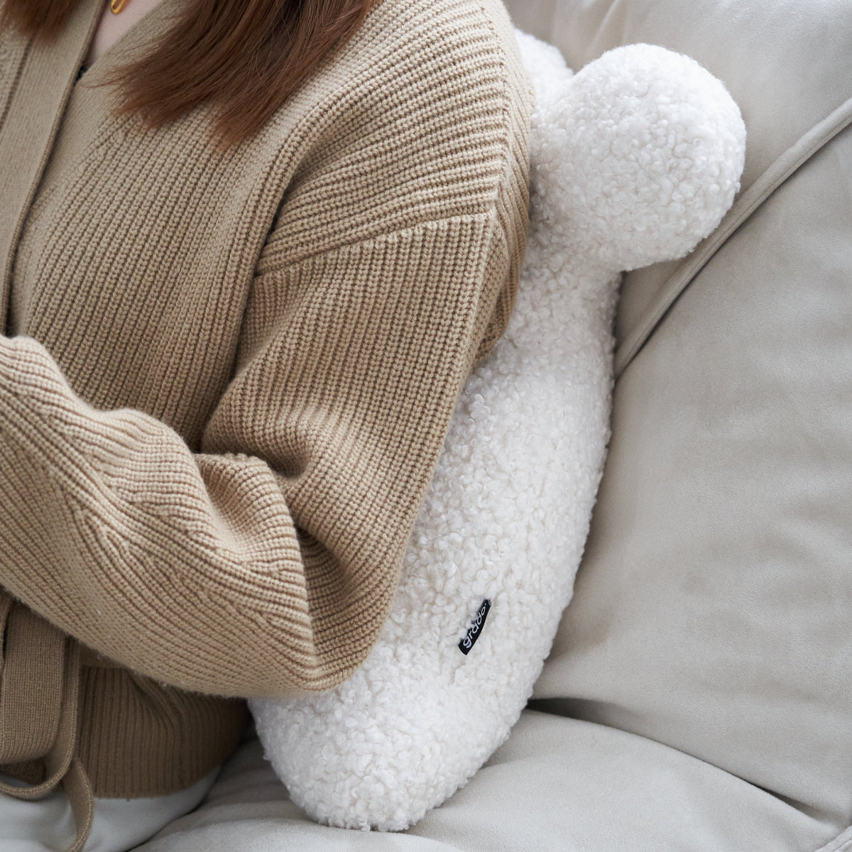 Person wearing a beige sweater sitting on a white couch with a fluffy white pillow.