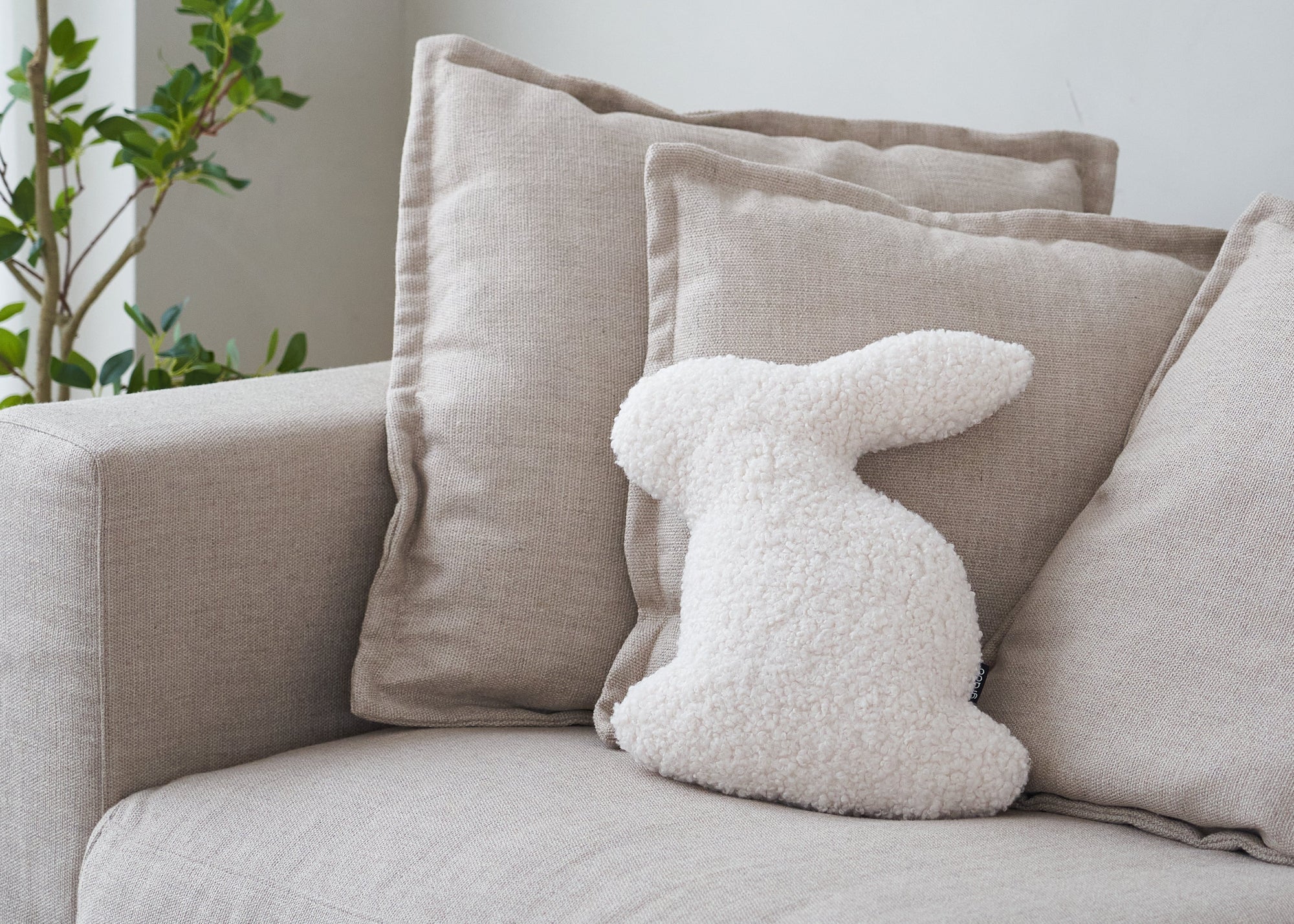 Beige sofa with decorative pillows and a fluffy white bunny toy, against a light gray wall with a plant.