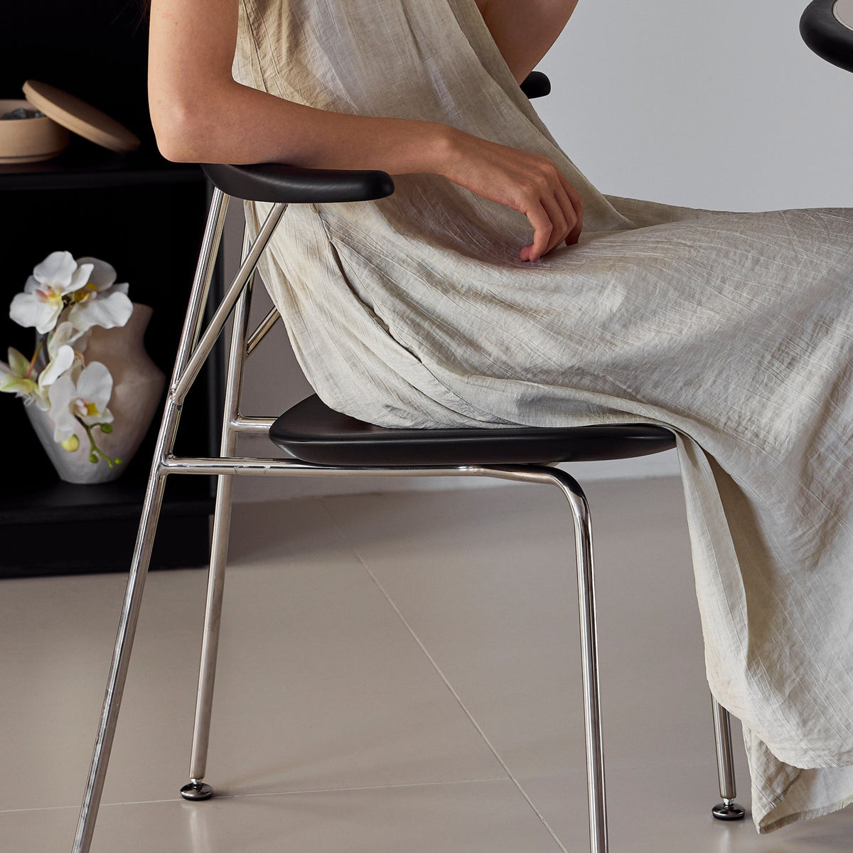 Woman in a light dress sitting on a chair, holding a small object in a room with a shelf and table.