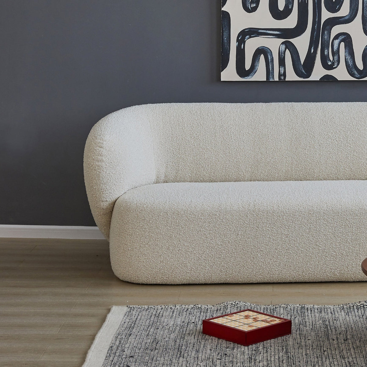 A large square abstract wall art print with black winding brushstrokes on a white background, displayed above a cream bouclé sofa in a modern living room setting.