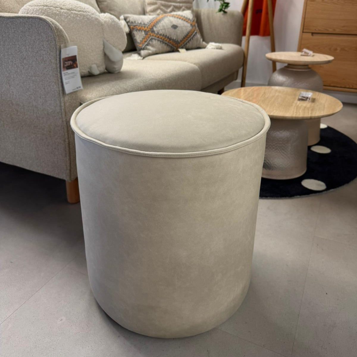 Beige ottoman in a living room setting with a sofa and small tables.